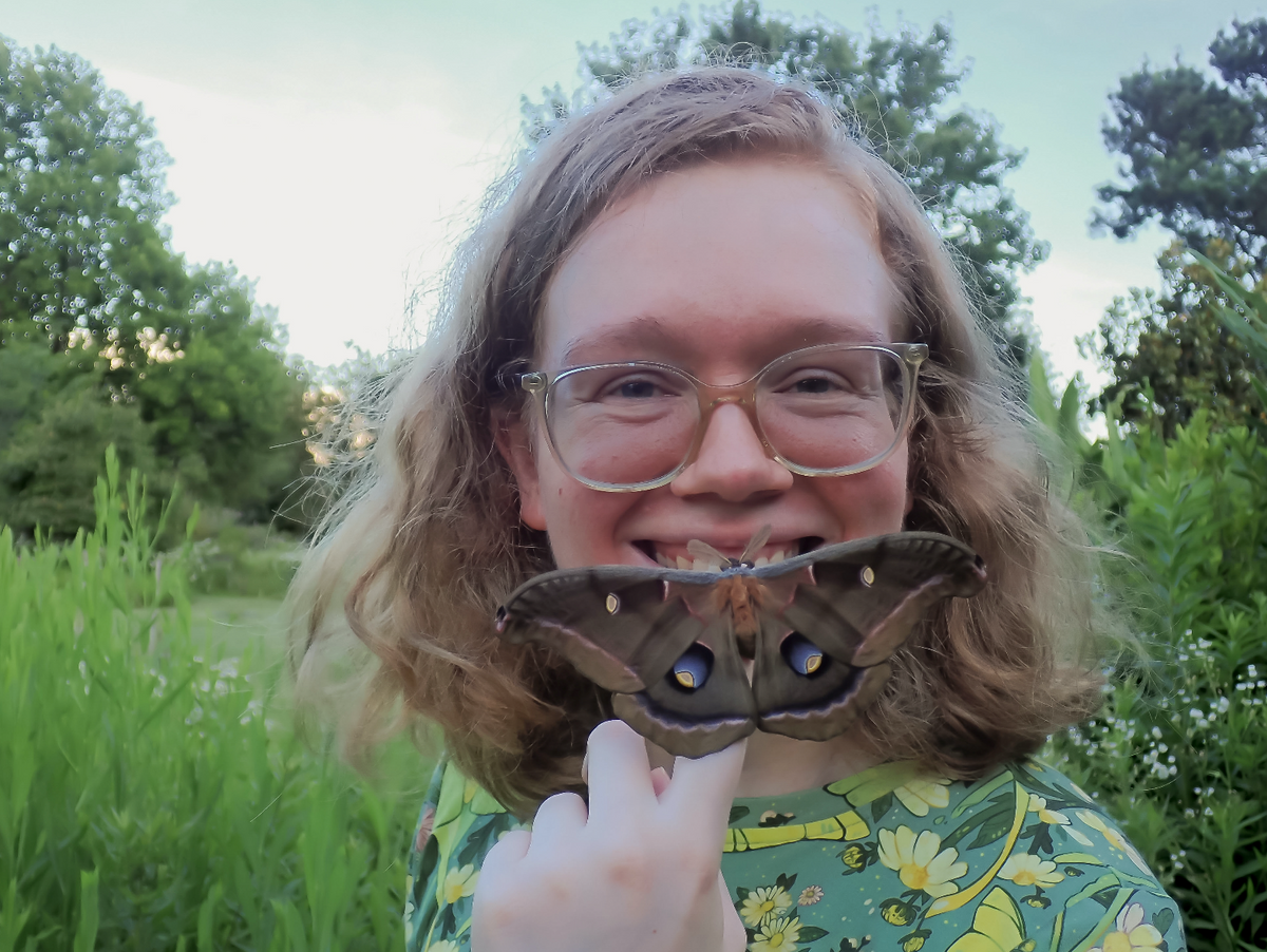 Allison Stoiser outdoors in a grassy field with trees in the background, wearing round eyeglasses and a green floral shirt, holding a Polyphemus moth near her face.