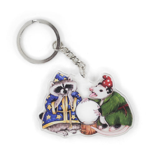 A keychain of a raccoon in starry blue wizard robes peering into a crystal ball with an opossum in green robes and a white spotted red hat on a white background.