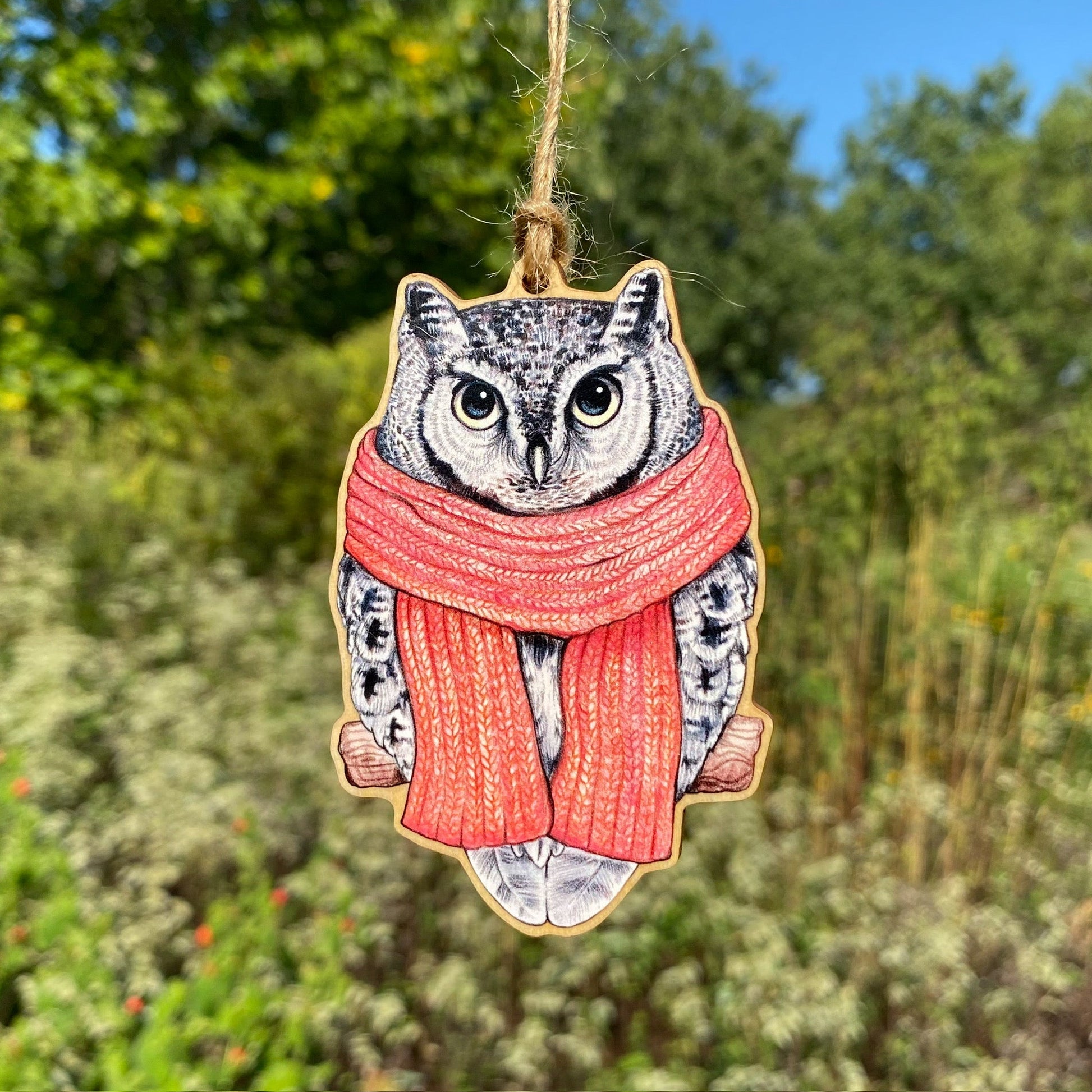 A wooden ornament strung with twine of a gray morph screech owl in a pink scarf.