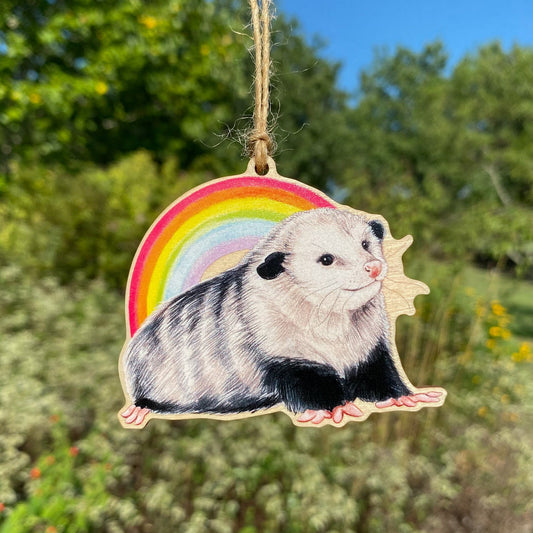 A wooden ornament strung with twine of a happy opossum laying in front of a rainbow.