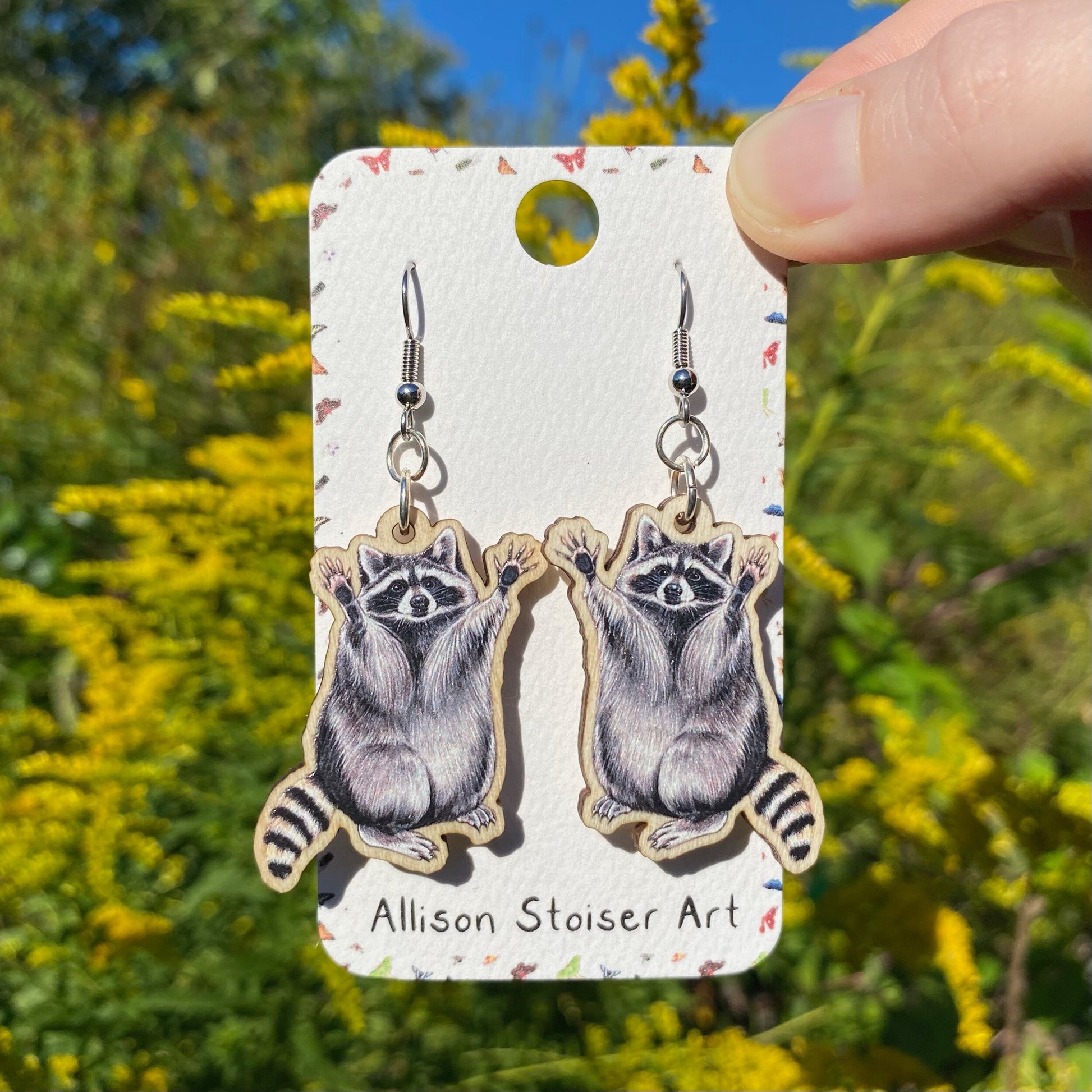 A hand holding a pair of wooden raccoon earrings.