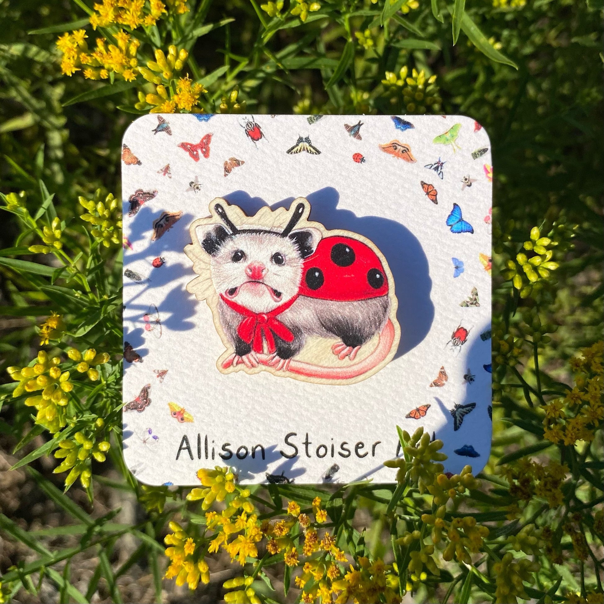 A sustainably sourced wooden pin featuring an opossum dressed as ladybug