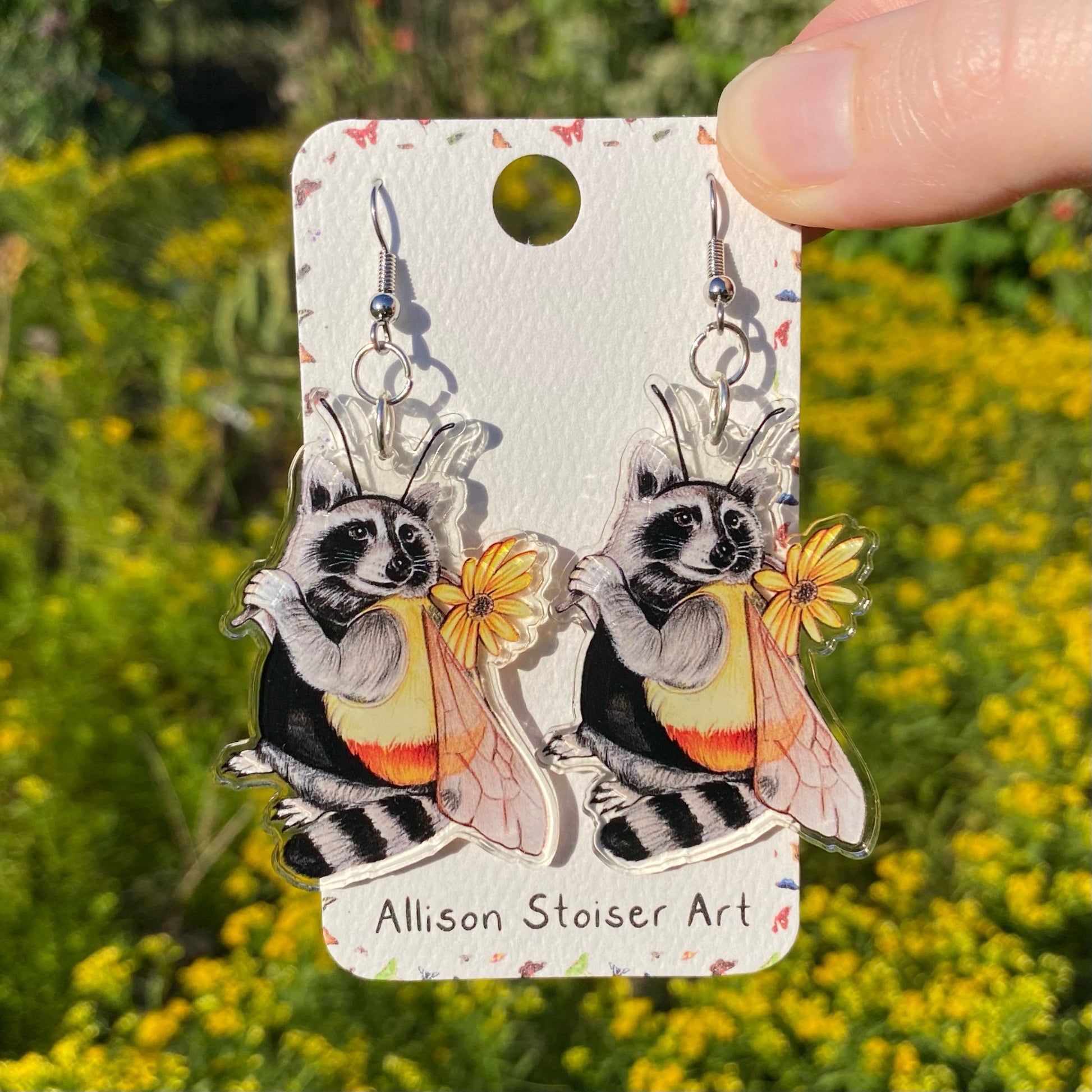 A hand holding a pair of acrylic earrings of a raccoon dressed as a bumblebee