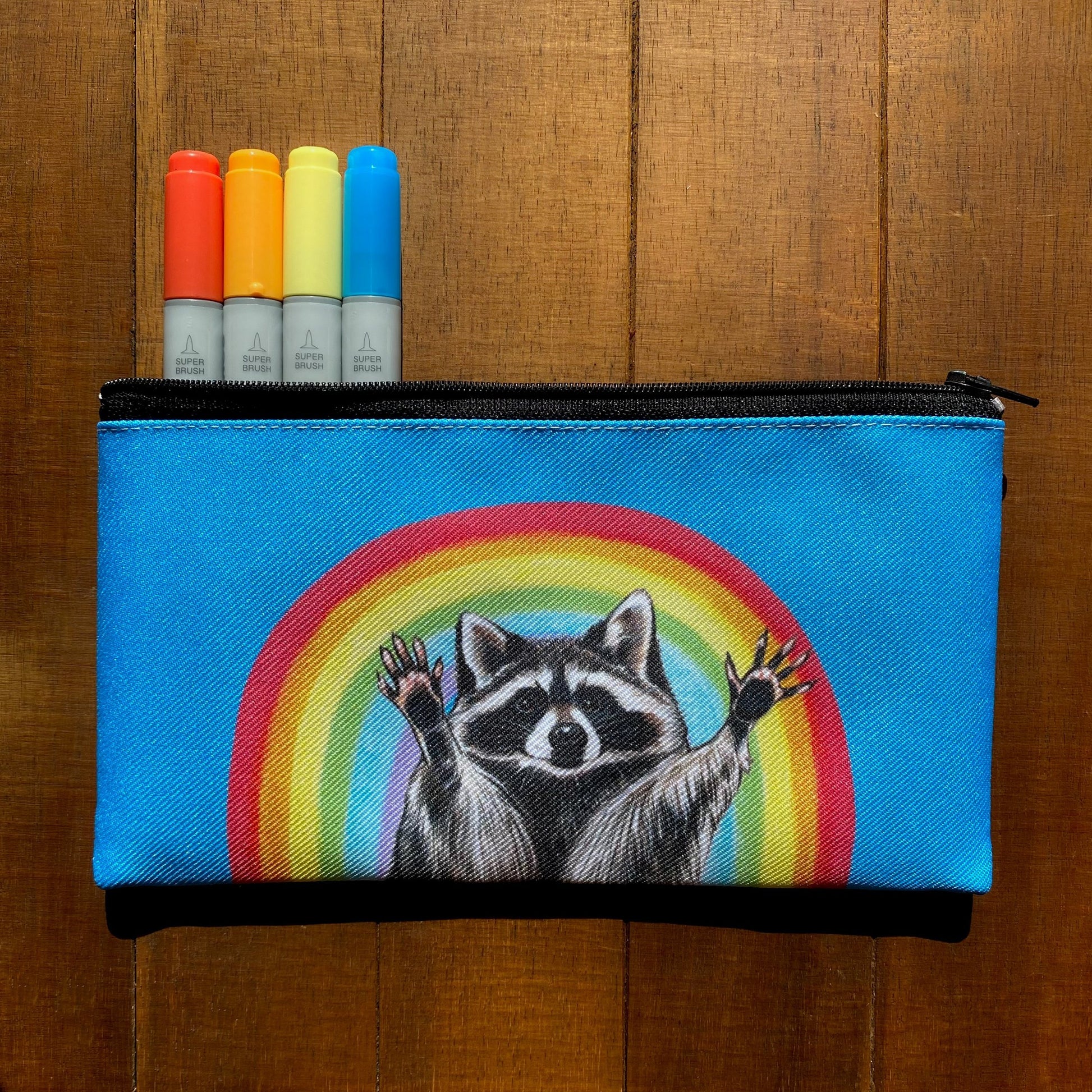 A zippered pouch featuring a raccoon with raised hands in front of a rainbow 