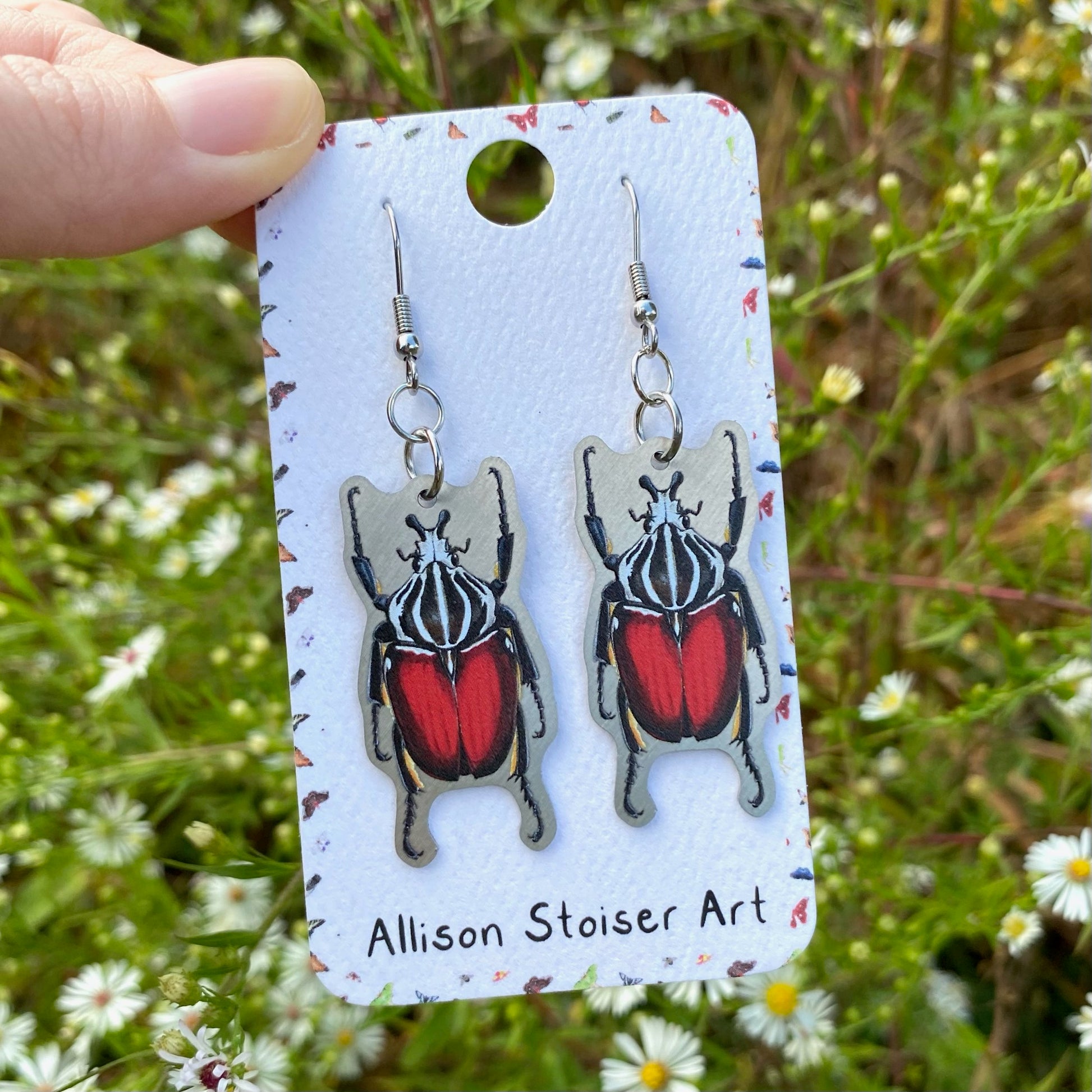 A hand holding a pair of metal goliath beetle earrings