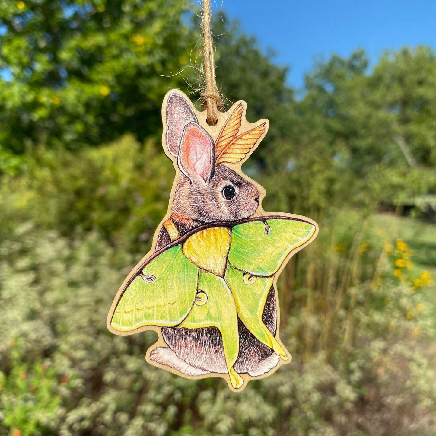 A wooden ornament strung with twine of an eastern cottontail rabbit dressed up as a luna moth.