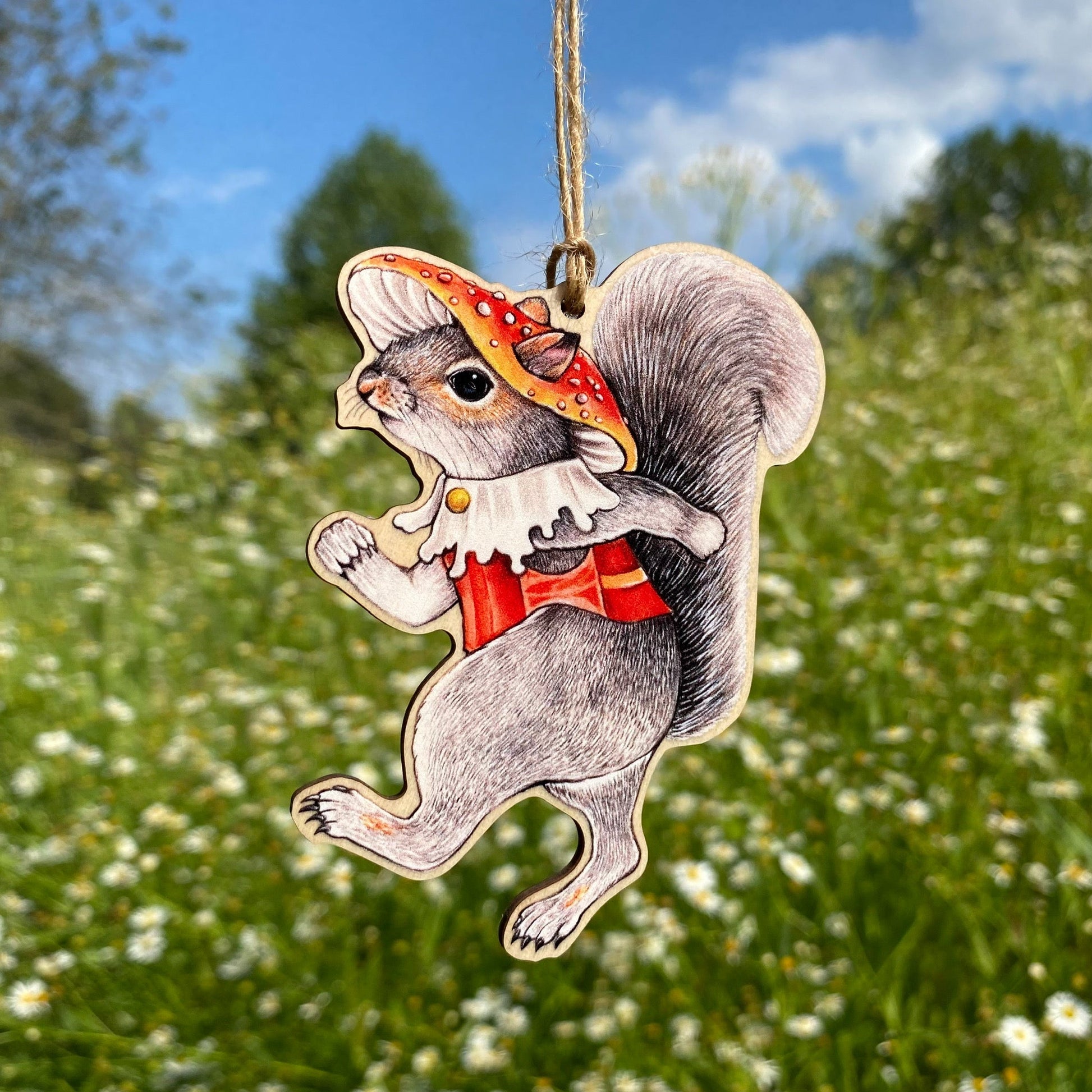 A wooden ornament strung with twine of a marching gray squirrel in a red toadstool hat and vest.