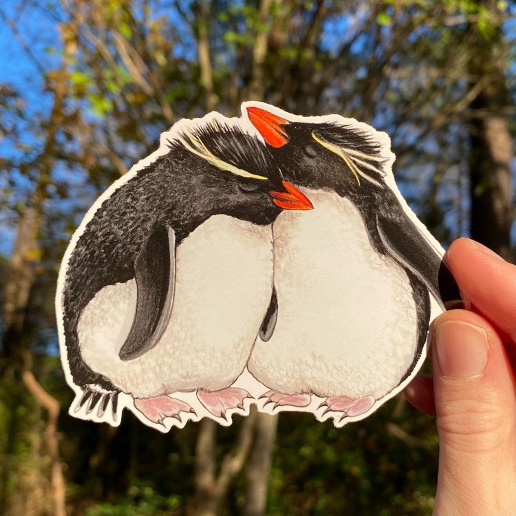 A hand holding a sticker featuring an illustration of a rockhopper penguin pair