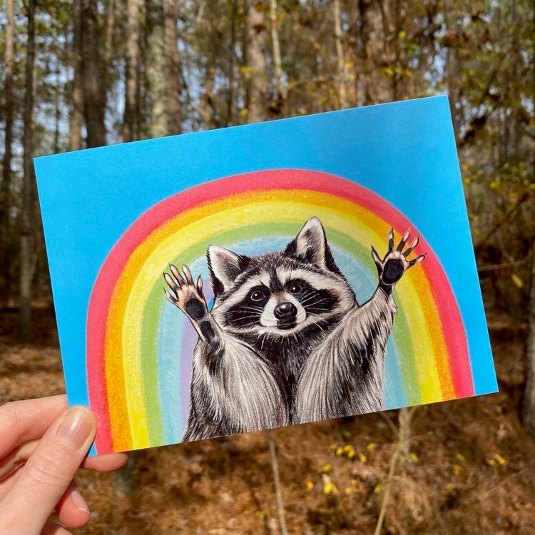 A postcard featuring a raccoon with his hands raised in front of a rainbow 