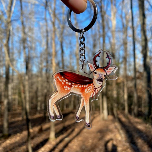 A keychain of a deer wearing stag beetle horns and a cloak being held in front of the forest.