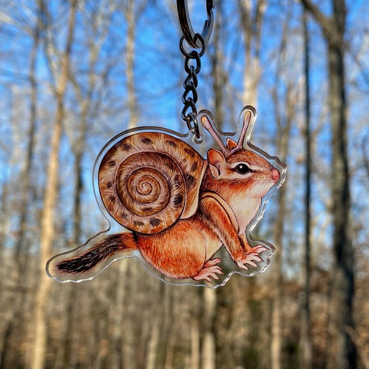 A keychain of a chipmunk in a snail shell being held in front of the forest.