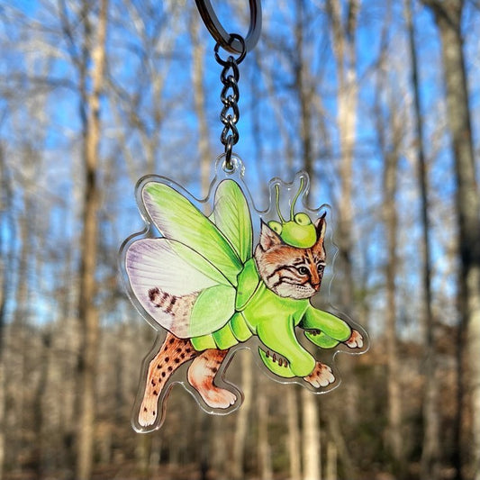 A keychain of a bobcat wearing a green praying mantis costume being held in front of the forest.