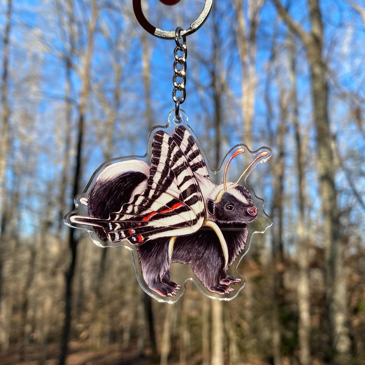 A keychain of a striped skunk in a zebra swallowtail costume being held in front of the forest.