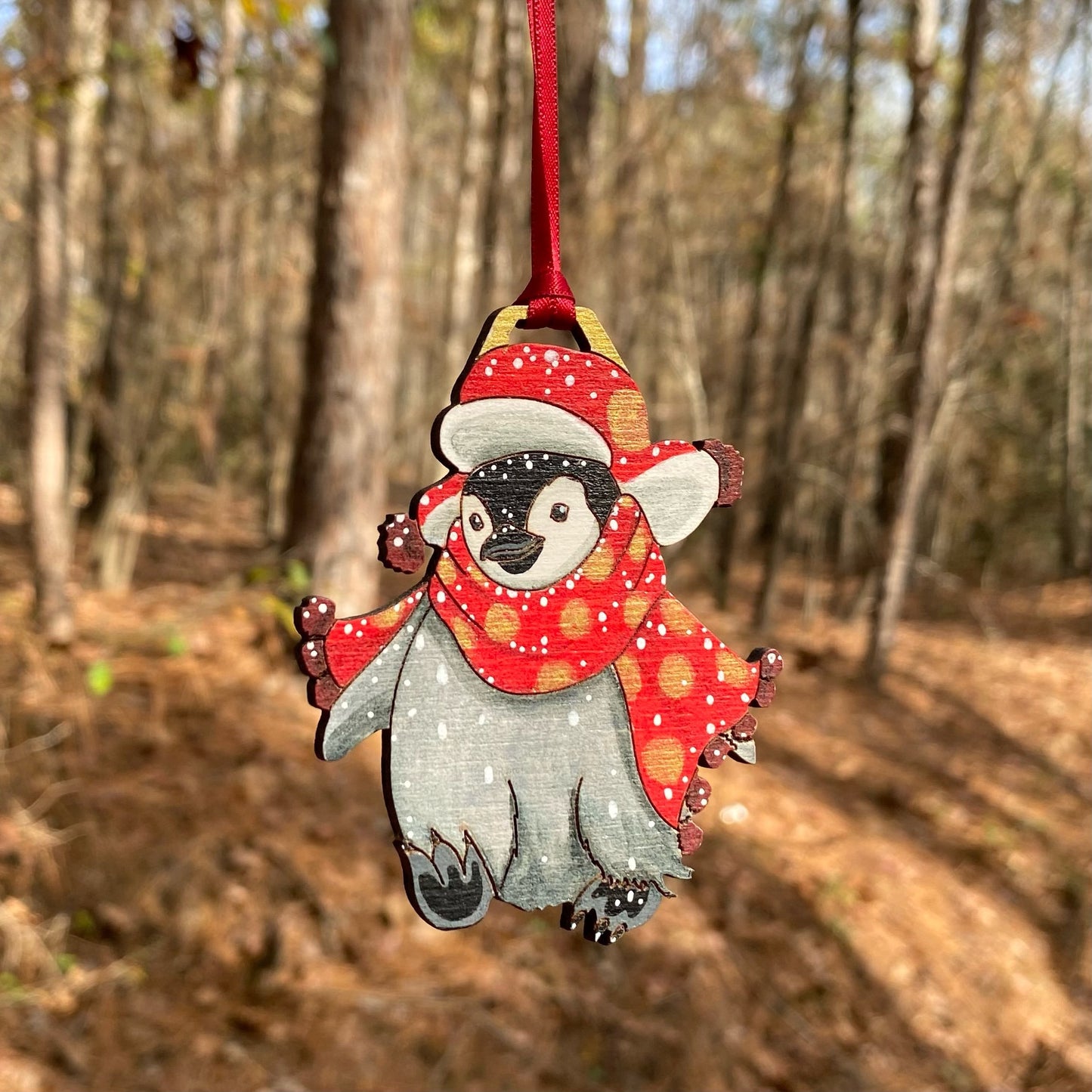 A hand-painted ornament of a penguin wearing a red scarf and hat with gold spots